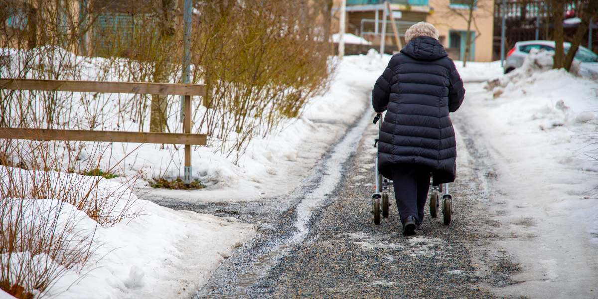 Elderly woman walking down snowy sidewalk with walker.